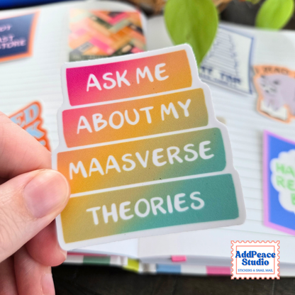 Close up of vinyl sticker in a rainbow color gradient book stack that says Ask me about my Maasverse theories, with AddPeace Studio stickers on an open notebook in the background.