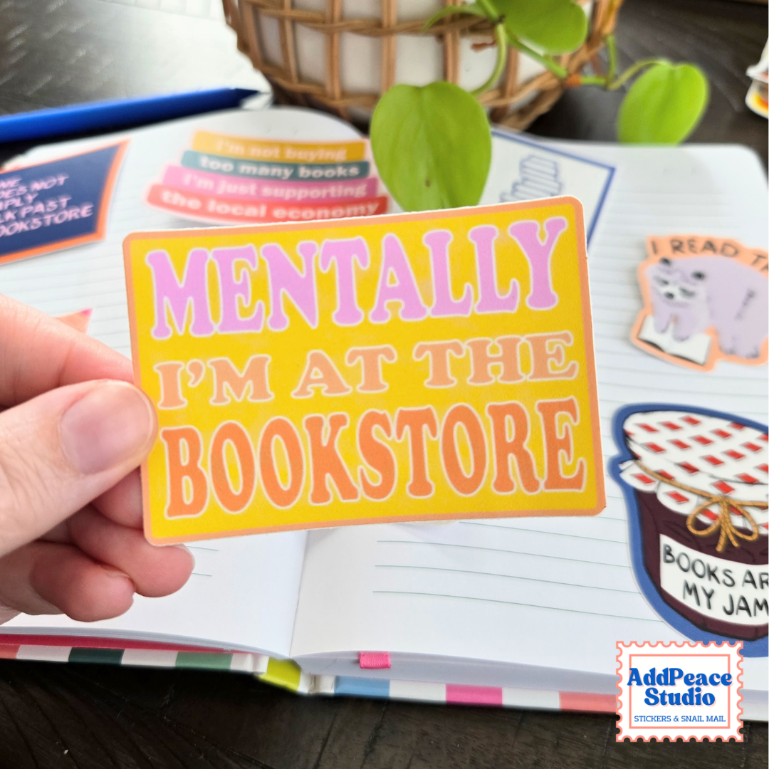 A hand holds a rectangular sticker that reads 'MENTALLY I'M AT THE BOOKSTORE' in pink and orange lettering on a yellow background. The sticker is shown over an open notebook filled with other colorful book-themed stickers, with a basket and green plant in the background.