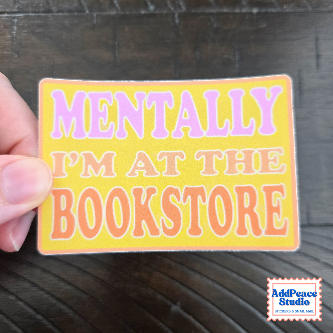 A hand holds a rectangular sticker with the text 'MENTALLY I'M AT THE BOOKSTORE' in pink and orange lettering on a yellow background, set against a dark wood surface. The AddPeace Studio logo is visible in the bottom right corner.