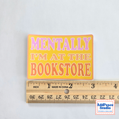 A rectangular sticker with the text 'MENTALLY I'M AT THE BOOKSTORE' in pink and orange lettering on a yellow background, shown above a wooden ruler for scale. A 'AddPeace Studio' watermark is visible in the bottom right corner.
