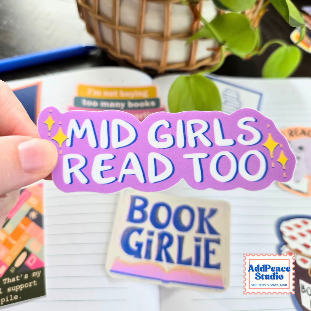A hand holds a die-cut sticker that reads 'MID GIRLS READ TOO' in white letters on a purple background with yellow sparkle accents. Other stickers, a notebook, and a plant are visible in the background.