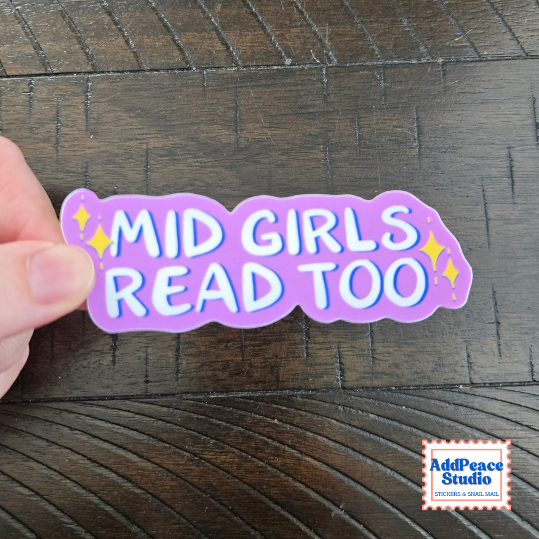 A hand holds a die-cut sticker that reads 'MID GIRLS READ TOO' in white letters with blue outlines on a purple background, with small yellow star accents, set against a dark wood surface. An 'AddPeace Studio' logo is visible in the bottom right corner.