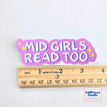 A die-cut sticker with the text 'MID GIRLS READ TOO' in white bubble letters on a pink background, shown above a wooden ruler for scale. The sticker has small yellow sparkle accents.