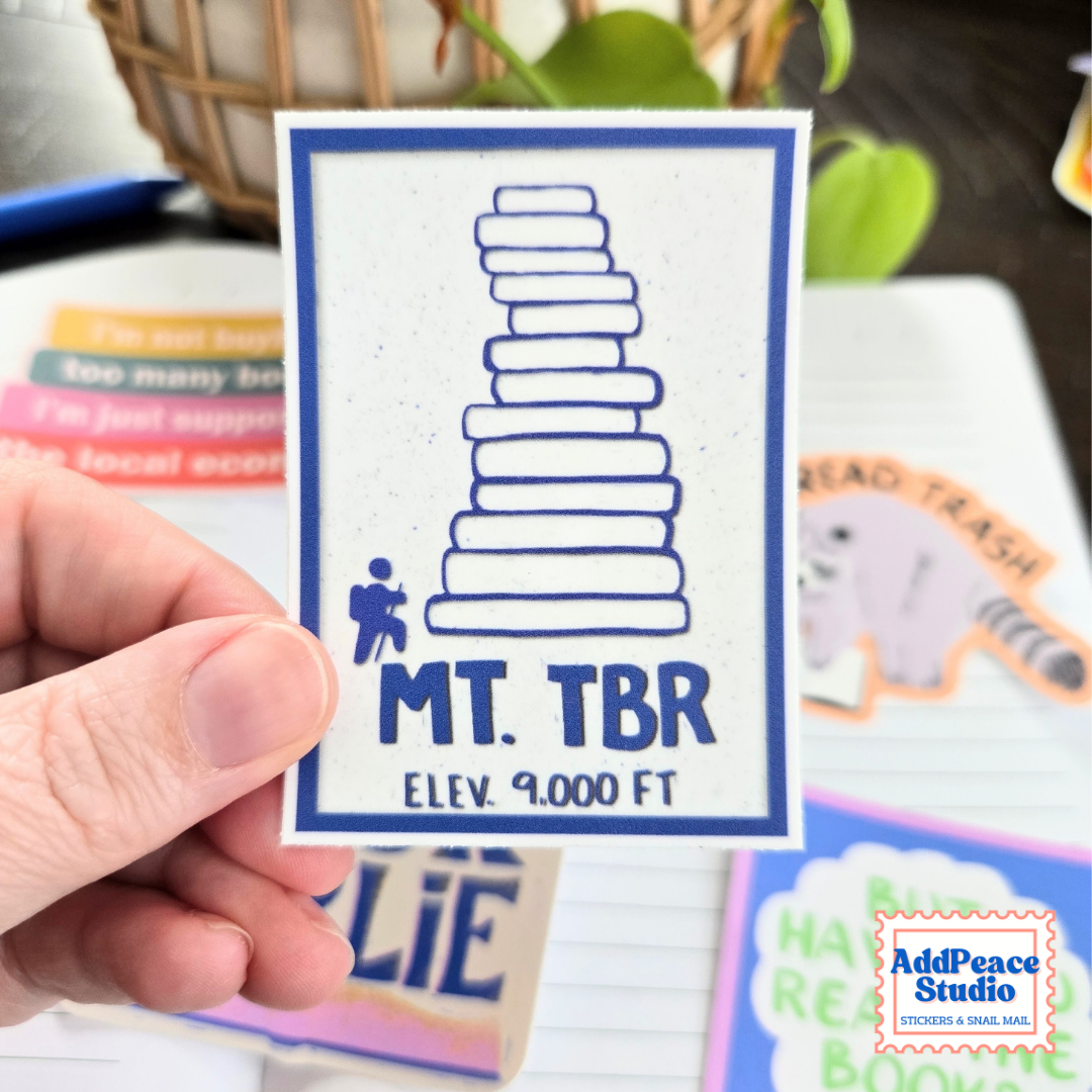A hand holds a square white sticker featuring a blue border, a blue stacked-book graphic, and the text 'MT. TBR ELEV. 9000 FT