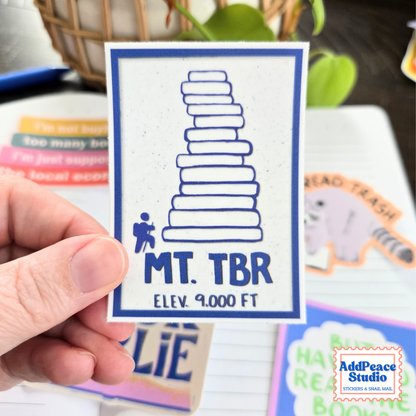 A hand holds a square white sticker featuring a blue border, a blue stacked-book graphic, and the text 'MT. TBR ELEV. 9000 FT