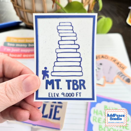 A hand holds a square white sticker featuring a blue border, a blue stacked-book graphic, and the text 'MT. TBR ELEV. 9000 FT