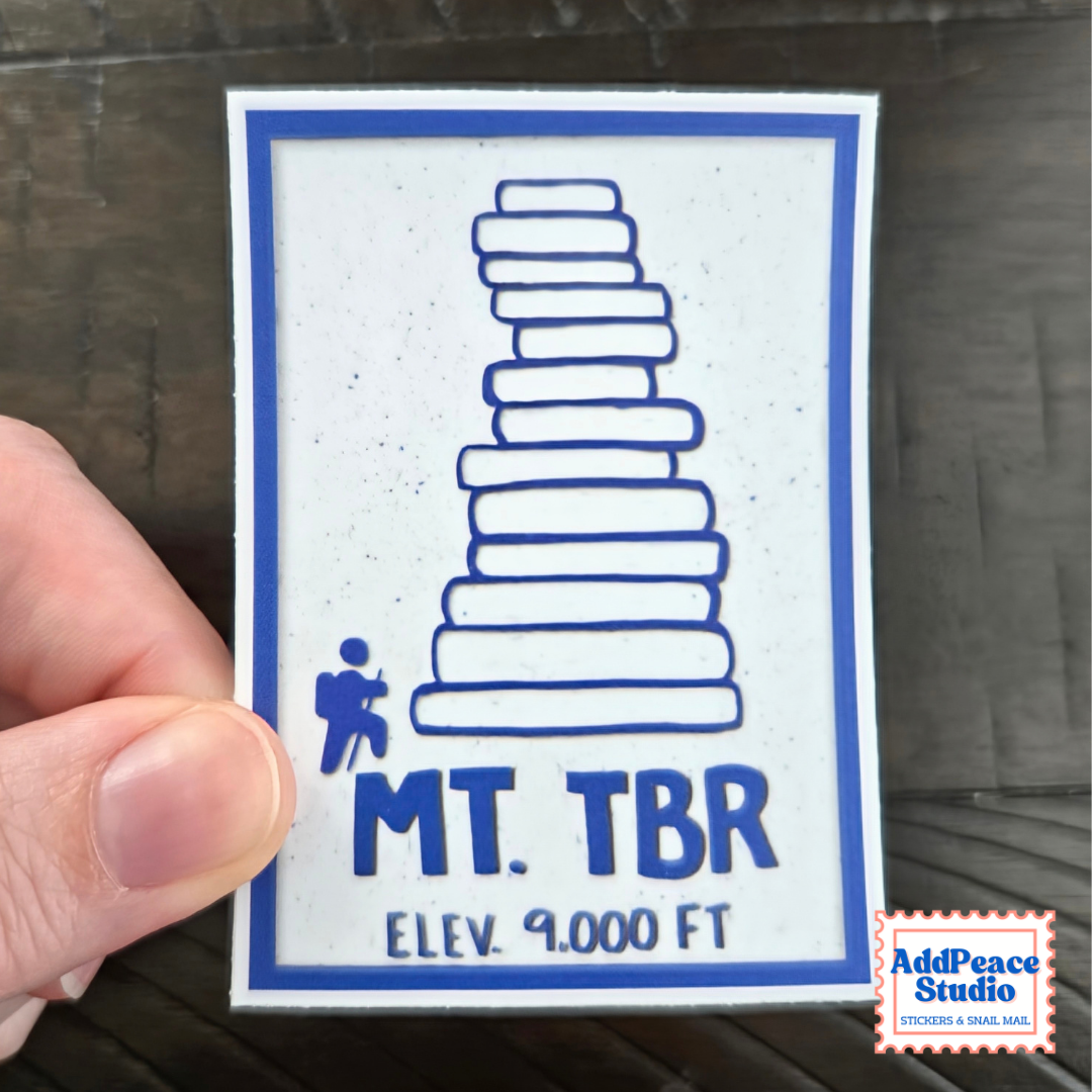 A hand holds a square white sticker with a blue border and blue printed graphic of a book stack, labeled 'MT. TBR' and 'ELEV. 9000 FT