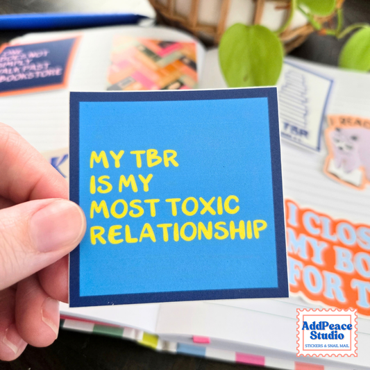 A hand holds a square blue sticker with yellow text that reads 'MY TBR IS MY MOST TOXIC RELATIONSHIP'. The background shows a blurred book and plant, with an 'AddPeace Studio' logo in the bottom right corner.