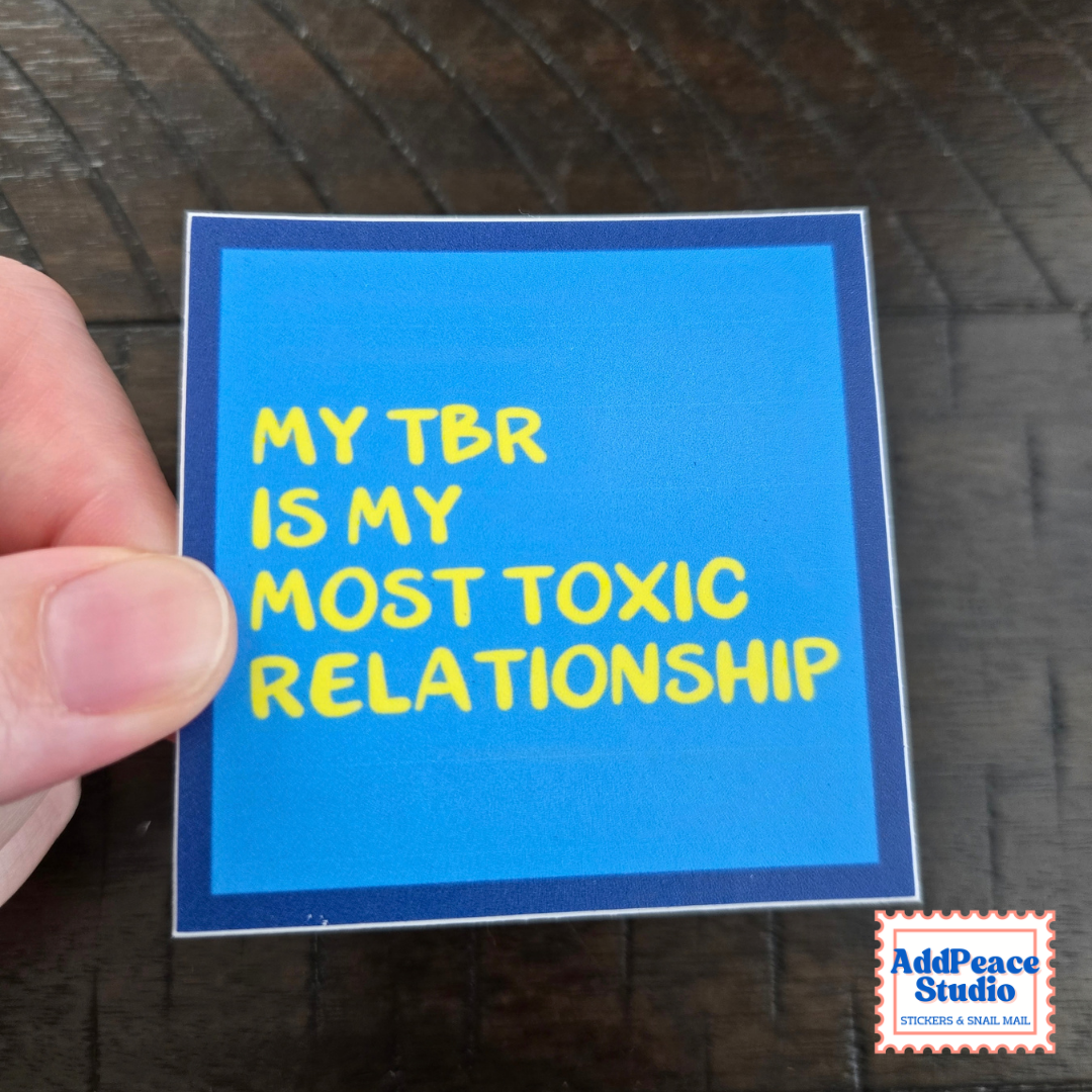 A hand holds a square blue sticker with yellow text that reads 'MY TBR IS MY MOST TOXIC RELATIONSHIP' against a dark wood background. An AddPeace Studio logo appears in the bottom right corner.