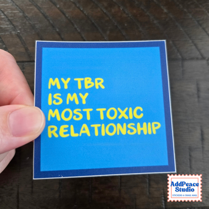 A hand holds a square blue sticker with yellow text that reads 'MY TBR IS MY MOST TOXIC RELATIONSHIP' against a dark wood background. An AddPeace Studio logo appears in the bottom right corner.