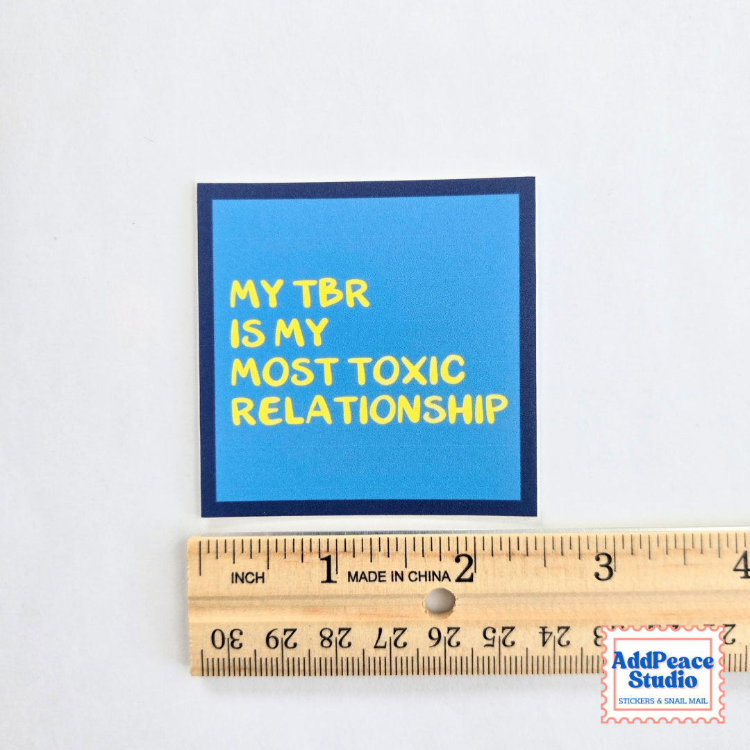 A square blue sticker with yellow text reading 'MY TBR IS MY MOST TOXIC RELATIONSHIP' and a dark navy border, shown above a wooden ruler for scale on a white background.