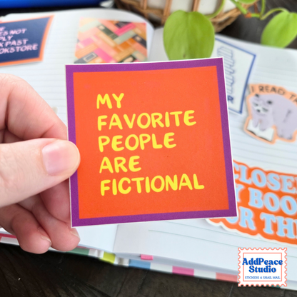 A hand holds a square orange sticker with a purple border and yellow text that reads 'MY FAVORITE PEOPLE ARE FICTIONAL'. The background shows an open notebook with colorful stickers and plant leaves.
