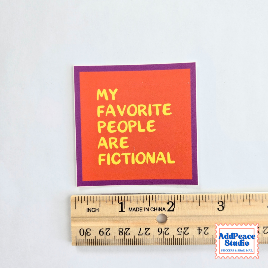 A square sticker with the text 'MY FAVORITE PEOPLE ARE FICTIONAL' in yellow on an orange-red background with a purple border, shown above a wooden ruler for scale on a white background.