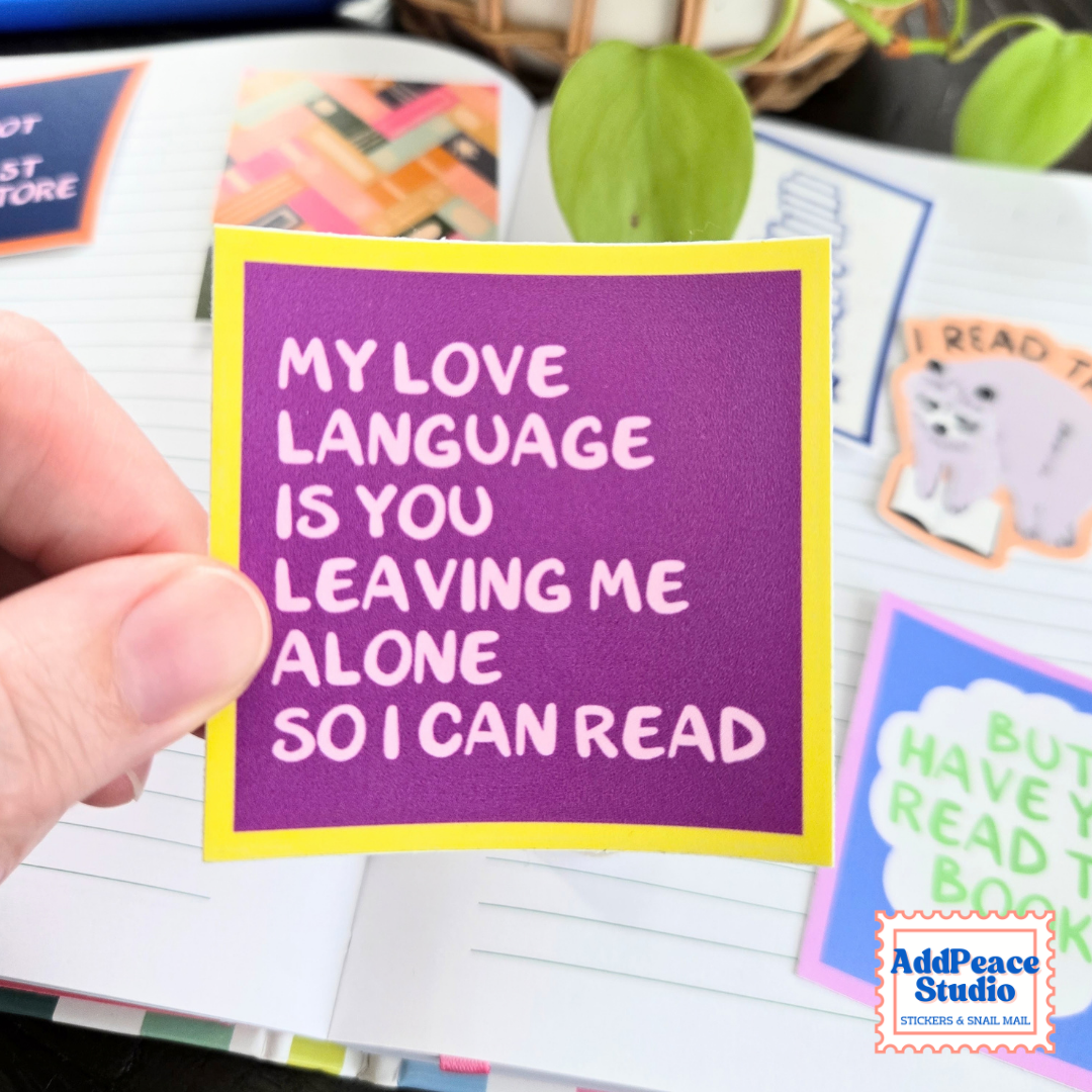 A hand holds a square purple sticker with a yellow border and white text that reads, 'MY LOVE LANGUAGE IS YOU LEAVING ME ALONE SO I CAN READ.' The sticker is shown over an open notebook with other colorful stickers visible in the background.