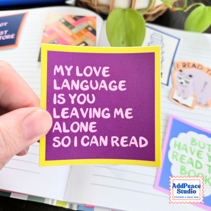 A hand holds a square purple sticker with a yellow border and white text that reads, 'MY LOVE LANGUAGE IS YOU LEAVING ME ALONE SO I CAN READ.' The sticker is shown over an open notebook with other colorful stickers visible in the background.
