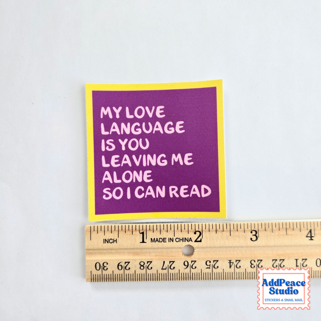 A square purple sticker with white text reading 'MY LOVE LANGUAGE IS YOU LEAVING ME ALONE SO I CAN READ', bordered in yellow, shown next to a wooden ruler on a white background. An AddPeace Studio watermark is in the bottom right corner.