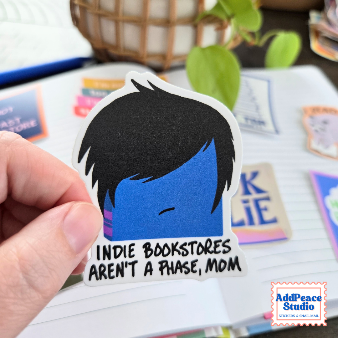 A hand holds a die-cut sticker featuring a minimalist blue and black face with the text 'INDIE BOOKSTORES AREN'T A PHASE, MOM'. The background shows a notebook, planner stickers, and a plant in a cozy desk setting.
