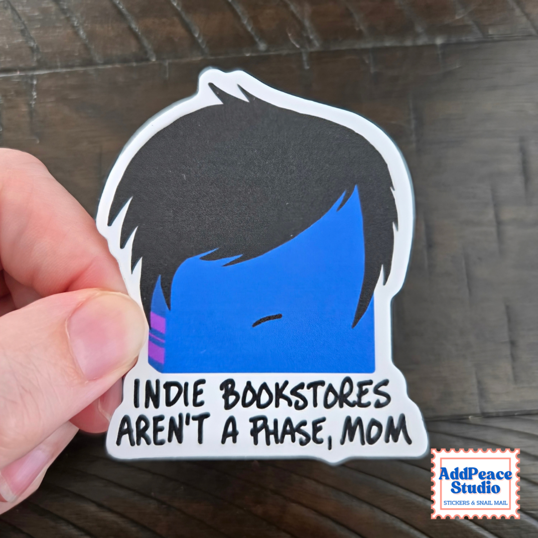 A hand holds a die-cut sticker featuring a stylized blue face with black hair and the text 'INDIE BOOKSTORES AREN'T A PHASE, MOM' on a dark wood background. An AddPeace Studio logo is visible in the bottom right corner.