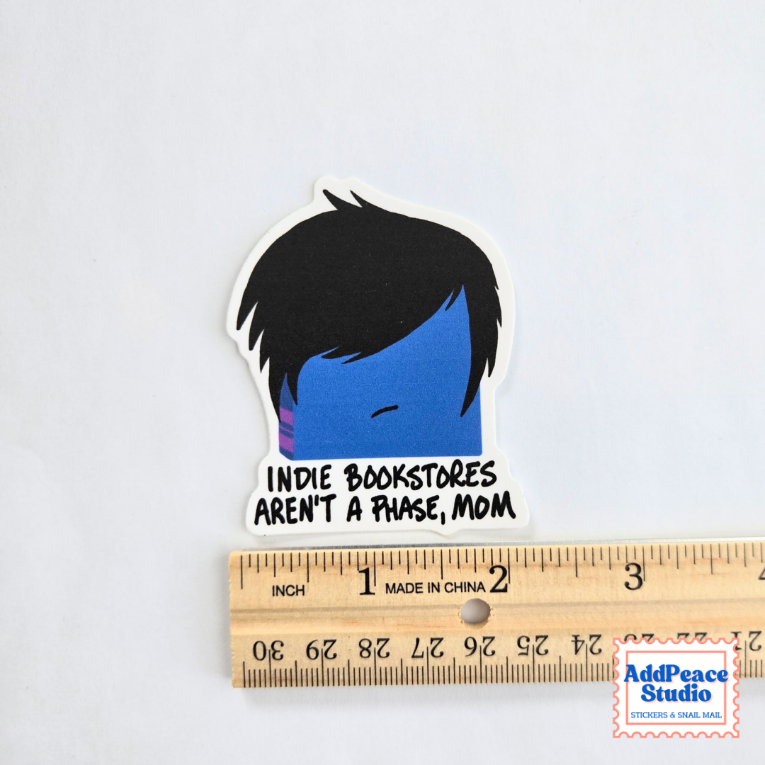 A die-cut sticker featuring a cartoon face with black hair, a blue face, and the text 'INDIE BOOKSTORES AREN'T A PHASE, MOM' in black. The sticker is shown above a wooden ruler for scale on a white background.