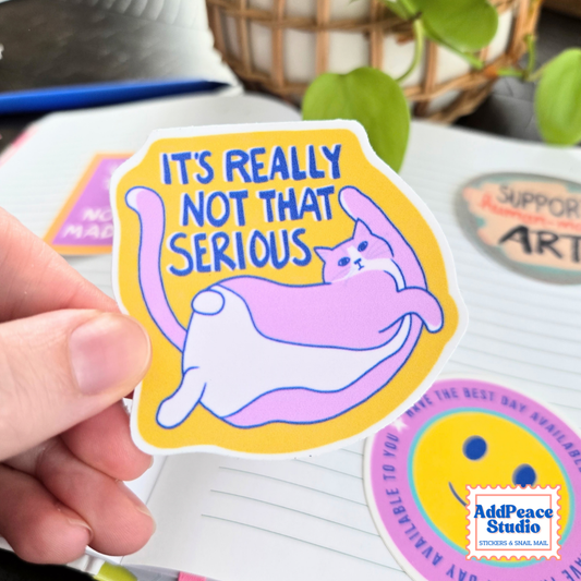 A hand holds a die-cut sticker featuring a pink and white cat lying on a yellow background with the text 'IT'S REALLY NOT THAT SERIOUS' in blue. The sticker is shown in a styled setting with other stickers and a notebook in the background.