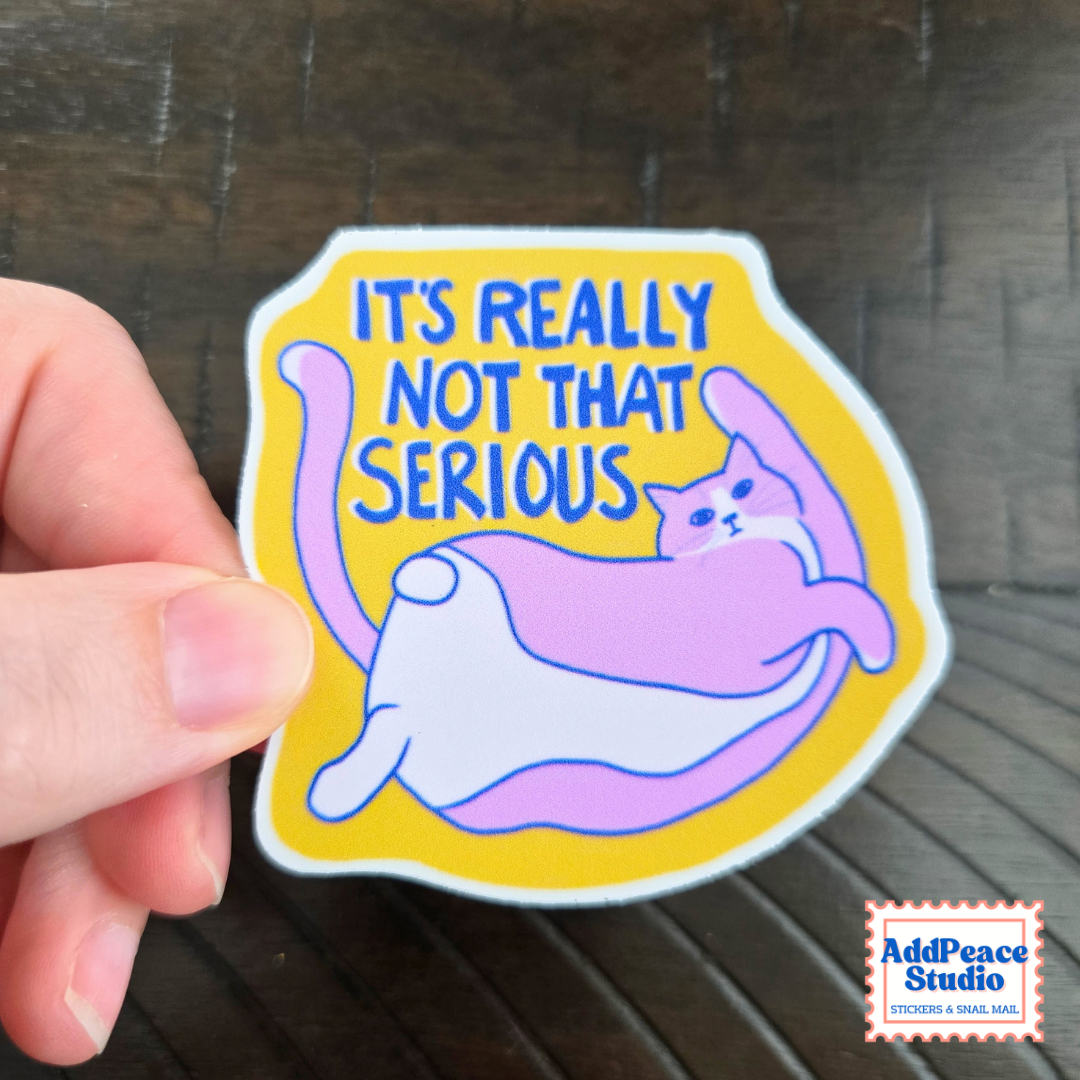 A hand holds a die-cut sticker featuring a cartoon cat lying on its side with the text 'IT'S REALLY NOT THAT SERIOUS' in blue on a yellow background. The sticker has a white border and is shown against a dark wood surface.