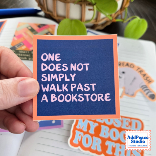 A hand holds a square sticker with a navy blue background and orange border, featuring the quote 'ONE DOES NOT SIMPLY WALK PAST A BOOKSTORE' in white text. The sticker is shown in a styled desk setting with notebooks, a plant, and other stickers.