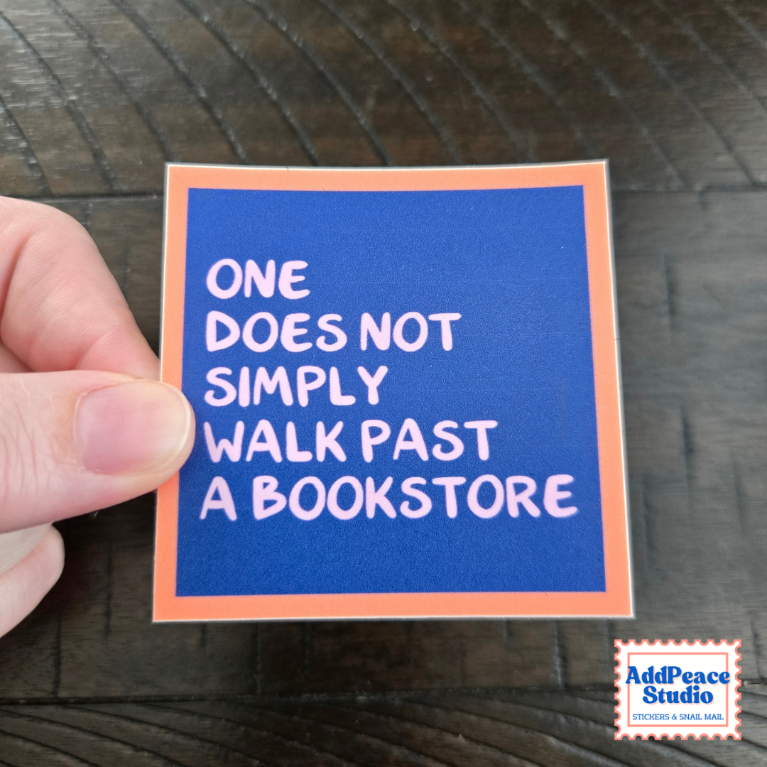 A hand holds a square sticker with a blue background and orange border, featuring the text 'ONE DOES NOT SIMPLY WALK PAST A BOOKSTORE' in white. The sticker is photographed on a dark wood surface, with an 'AddPeace Studio' logo in the bottom right corner.