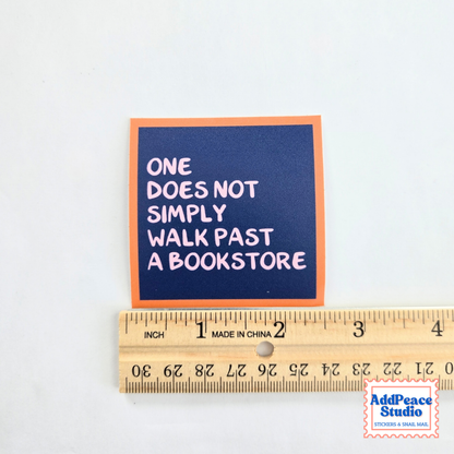 A square sticker with the quote 'ONE DOES NOT SIMPLY WALK PAST A BOOKSTORE' in white text on a dark blue background with an orange border, shown above a wooden ruler for scale on a white background.