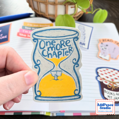 A hand holds a hand-drawn sticker of an hourglass with a hand holding a book popping out of the sand with the text One more chapter.