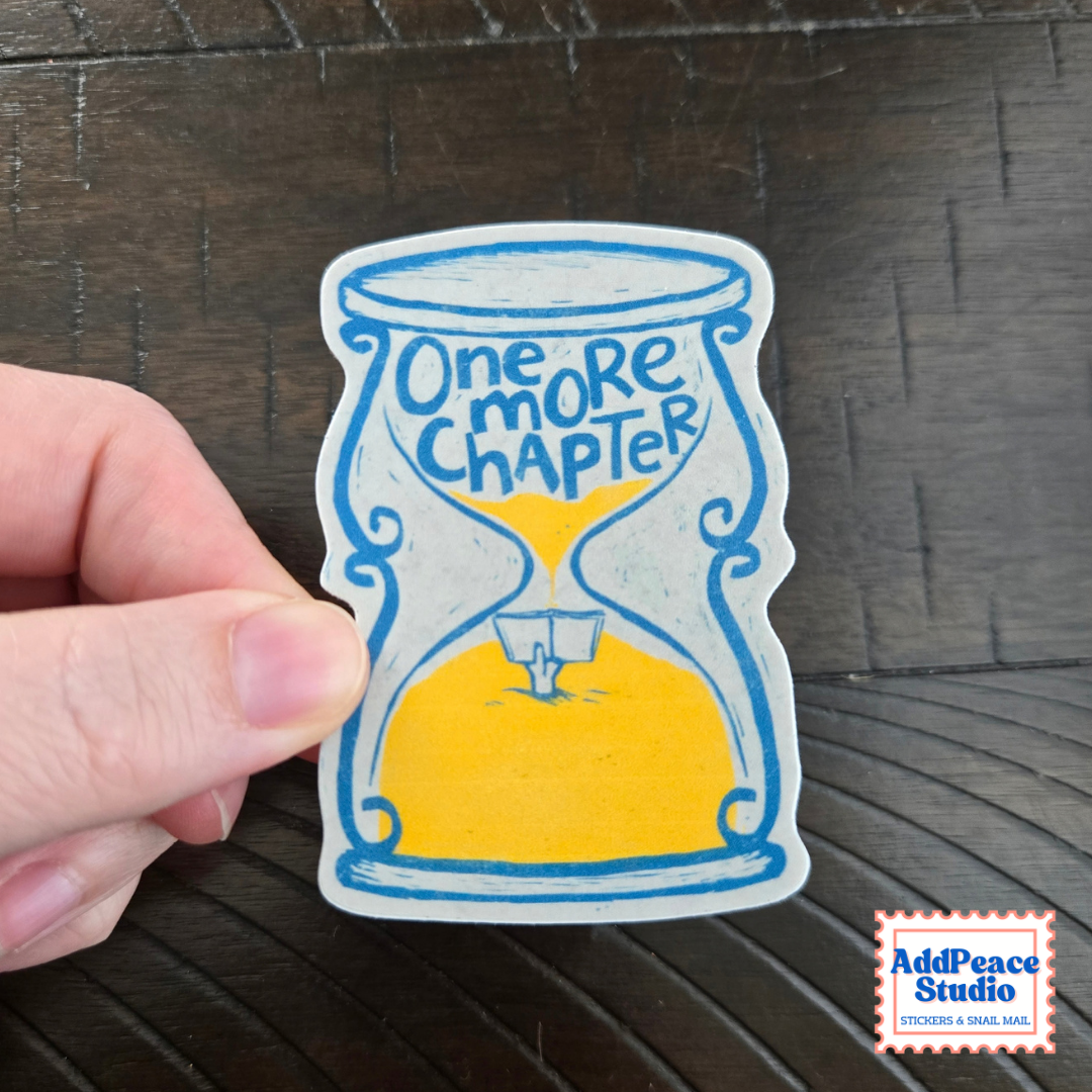 A hand holds a die-cut sticker featuring a hand-drawn hourglass with blue outlines, yellow sand, and the text 'One MORE CHAPTER' on a dark wood background. A 'AddPeace Studio' logo is visible in the bottom right corner.