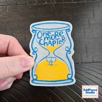 A hand holds a die-cut sticker featuring a hand-drawn hourglass with blue outlines, yellow sand, and the text 'One MORE CHAPTER' on a dark wood background. A 'AddPeace Studio' logo is visible in the bottom right corner.