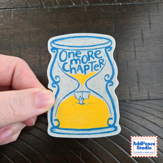 A hand holds a die-cut sticker featuring a hand-drawn hourglass with blue outlines, yellow sand, and the text 'One MORE CHAPTER' on a dark wood background. A 'AddPeace Studio' logo is visible in the bottom right corner.