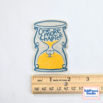 A die-cut sticker featuring an hourglass with yellow sand and the text 'One more chapter' in blue, shown above a wooden ruler for scale on a white background.