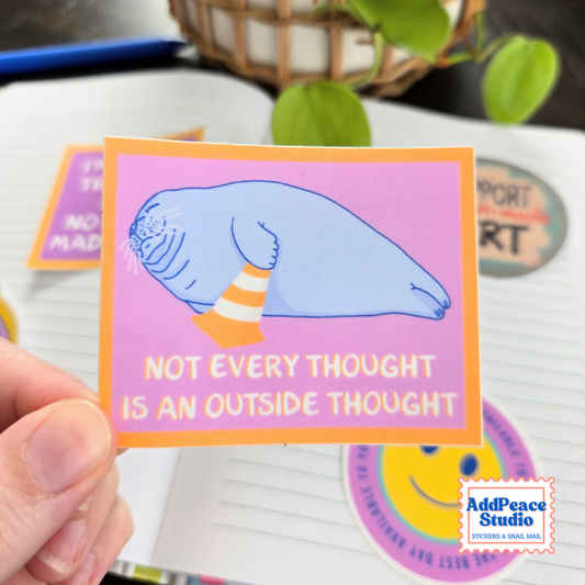 A hand holds a rectangular sticker with a pink background, orange border, and a blue seal illustration sitting in an orange-and-white striped cone. The sticker text reads, "NOT EVERY THOUGHT IS AN OUTSIDE THOUGHT.