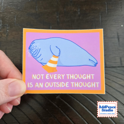 A hand holds a rectangular sticker featuring a blue seal on a pink background with an orange border and the text 'NOT EVERY THOUGHT IS AN OUTSIDE THOUGHT'. The sticker is photographed over a dark wood surface, with an AddPeace Studio logo in the bottom right corner.