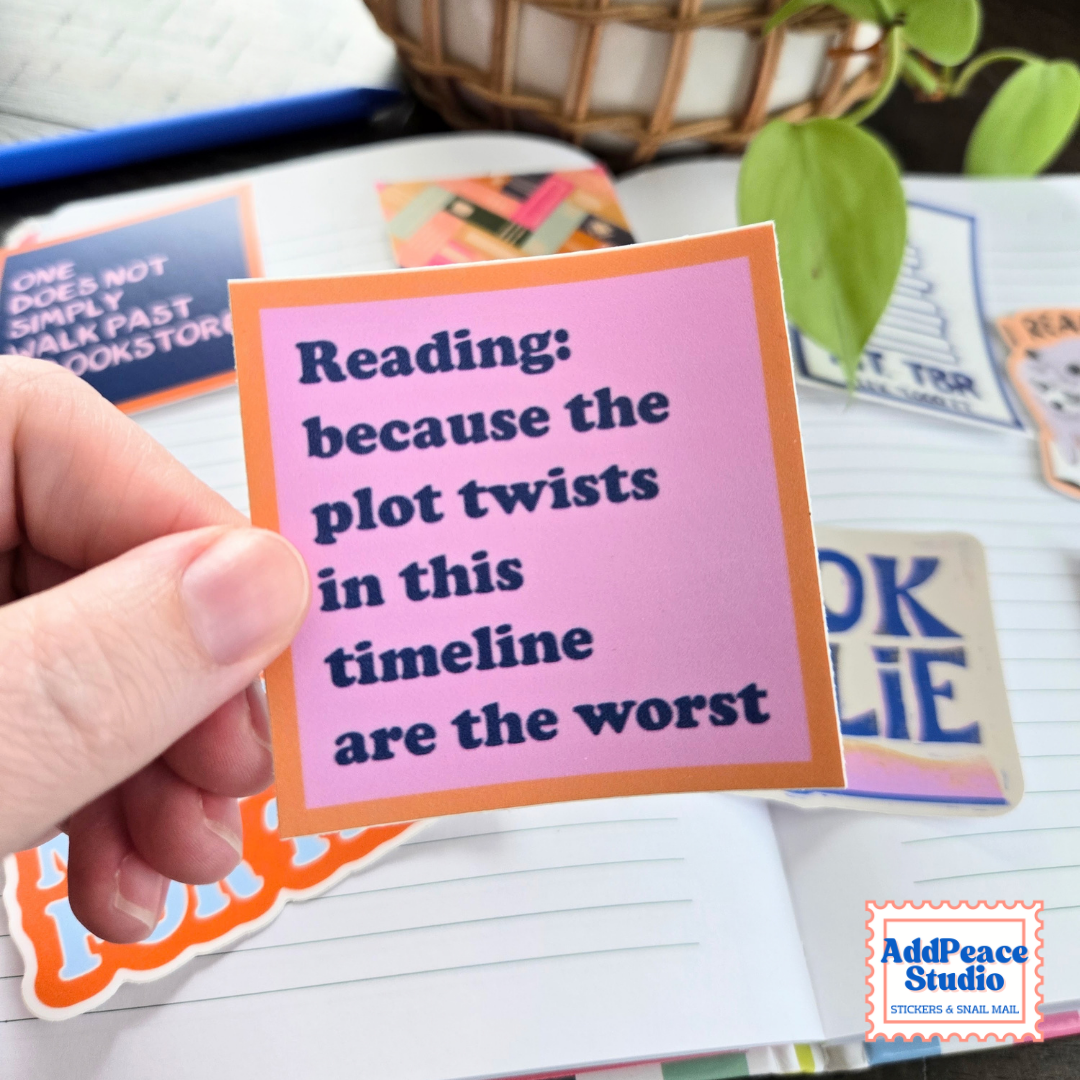 A hand holds a square sticker with a pink background and orange border, featuring the text 'Reading: because the plot twists in this timeline are the worst' in dark blue lettering. Other colorful stickers and a lined notebook are visible in the background, with a small 'AddPeace Studio' logo in the bottom right corner.