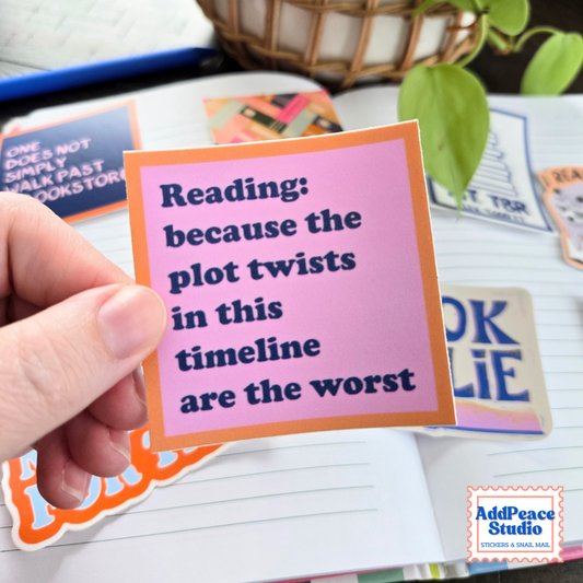 A hand holds a square sticker with a pink background and orange border, featuring the text 'Reading: because the plot twists in this timeline are the worst' in dark blue lettering. Other colorful stickers and a lined notebook are visible in the background, with a small 'AddPeace Studio' logo in the bottom right corner.