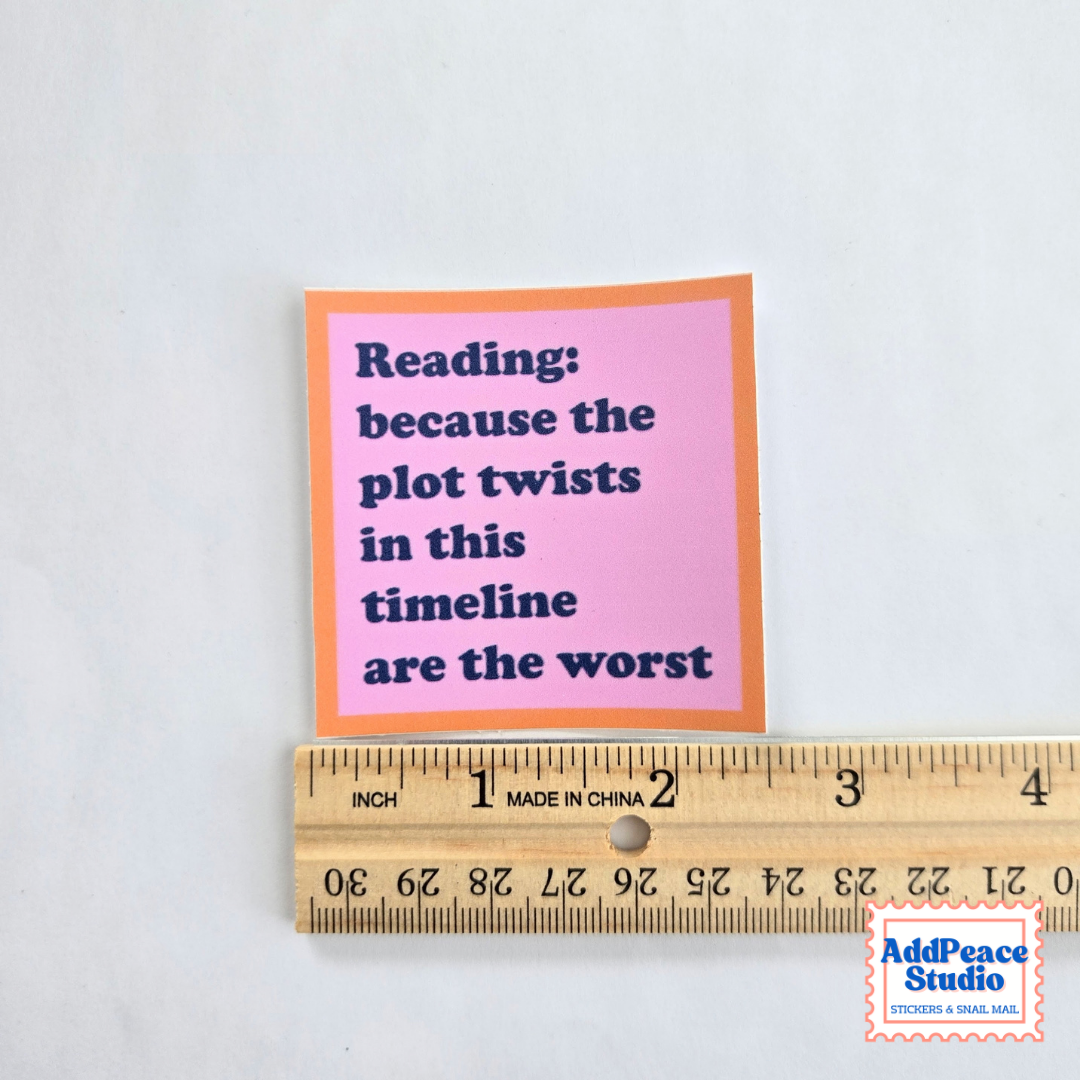 A square pink and orange sticker with dark blue text reading 'Reading: because the plot twists in this timeline are the worst' shown next to a wooden ruler for scale on a white background.