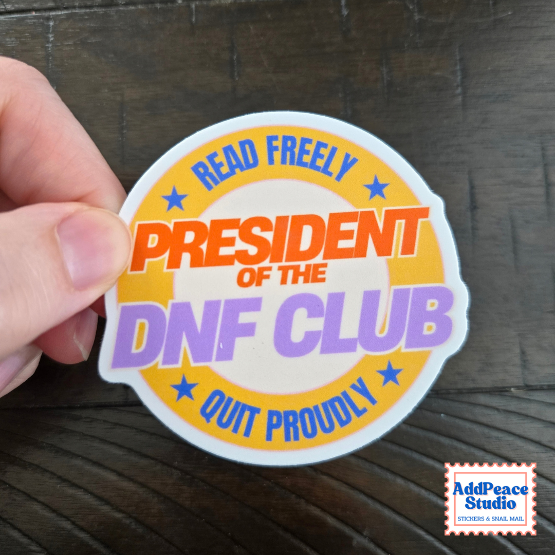 A hand holds a round sticker with the text 'READ FREELY PRESIDENT OF THE DNF CLUB QUIT PROUDLY' in orange, purple, and blue lettering on a white background, set against a dark wood surface. An 'AddPeace Studio' watermark is visible in the bottom right corner.