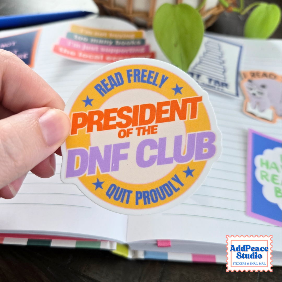 A hand holds a die cut sticker that says President of the DNF Club, Read Freely, Quit Proudly