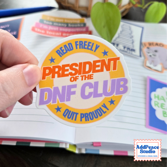 A hand holds a die cut sticker that says President of the DNF Club, Read Freely, Quit Proudly
