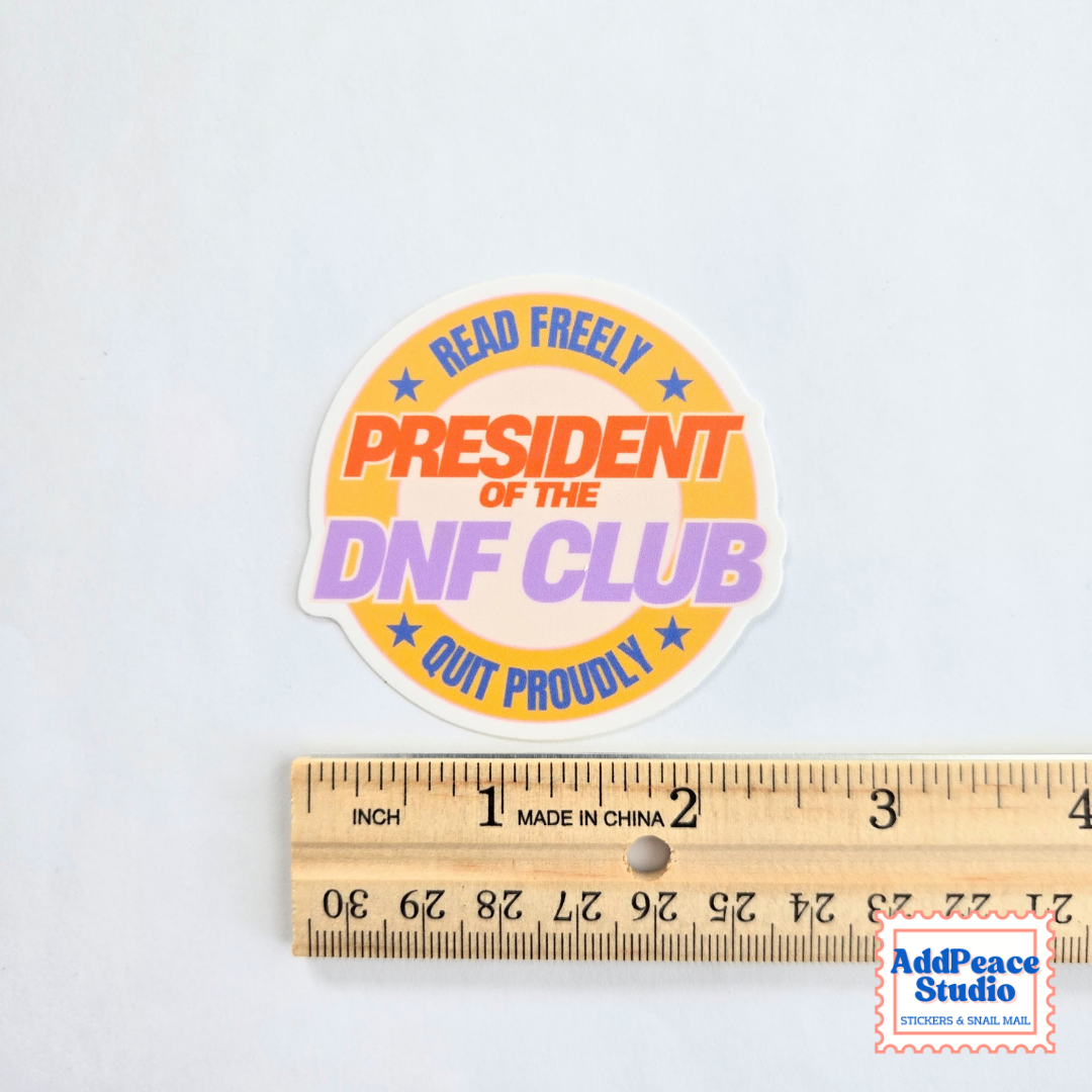 A round sticker with the text 'READ FREELY PRESIDENT OF THE DNF CLUB QUIT PROUDLY' in blue, orange, and purple lettering on a white background, shown above a wooden ruler for scale. A 'AddPeace Studio' watermark is in the bottom right corner.