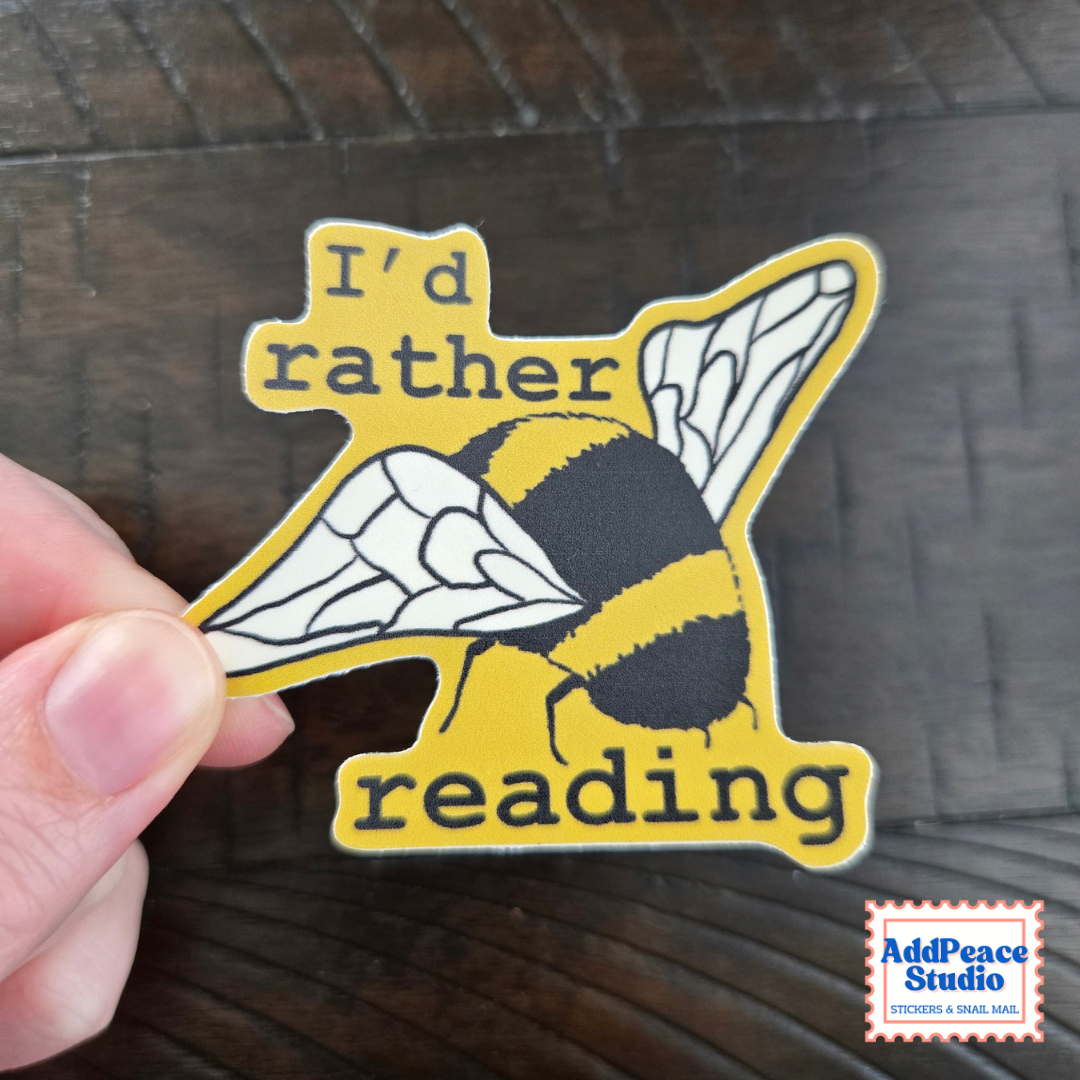A hand holds a die-cut sticker featuring a yellow and black bumblebee with the text 'I'd rather reading' in dark lettering, set against a dark wood background. An 'AddPeace Studio' brand logo is visible in the bottom right corner.