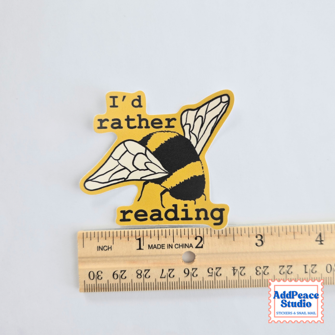 A die-cut sticker featuring a yellow and black bumblebee with the text 'I'd rather reading' in black, shown above a wooden ruler for scale on a white background.