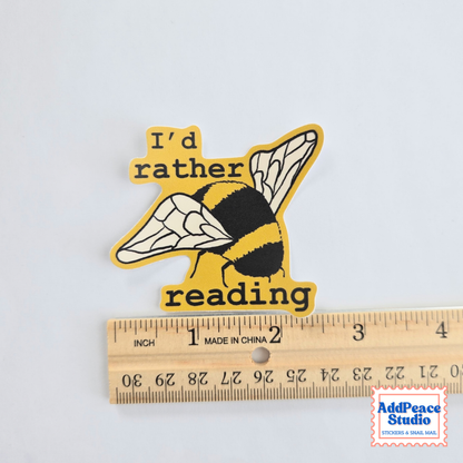 A die-cut sticker featuring a yellow and black bumblebee with the text 'I'd rather reading' in black, shown above a wooden ruler for scale on a white background.