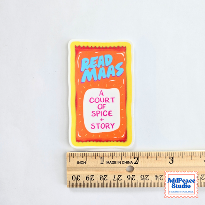 A rectangular sticker featuring a stylized orange and taco sauce packet design with the text 'READ MAAS' in blue and 'A COURT OF SPICE + STORY' in pink, shown above a wooden ruler for scale on a white background.