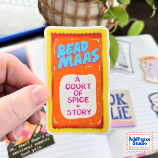 A hand holds a colorful sticker shaped like a taco sauce packet that reads 'READ MAAS' and 'A COURT OF SPICE + STORY'. The background is a blurred desk with other stickers and a plant.