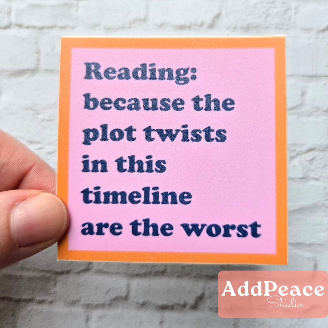 Hand holding a pink sticker with blue text “Reading because the plot twists in this timeline are the worst” featuring the brand 'AddPeace Studios'.