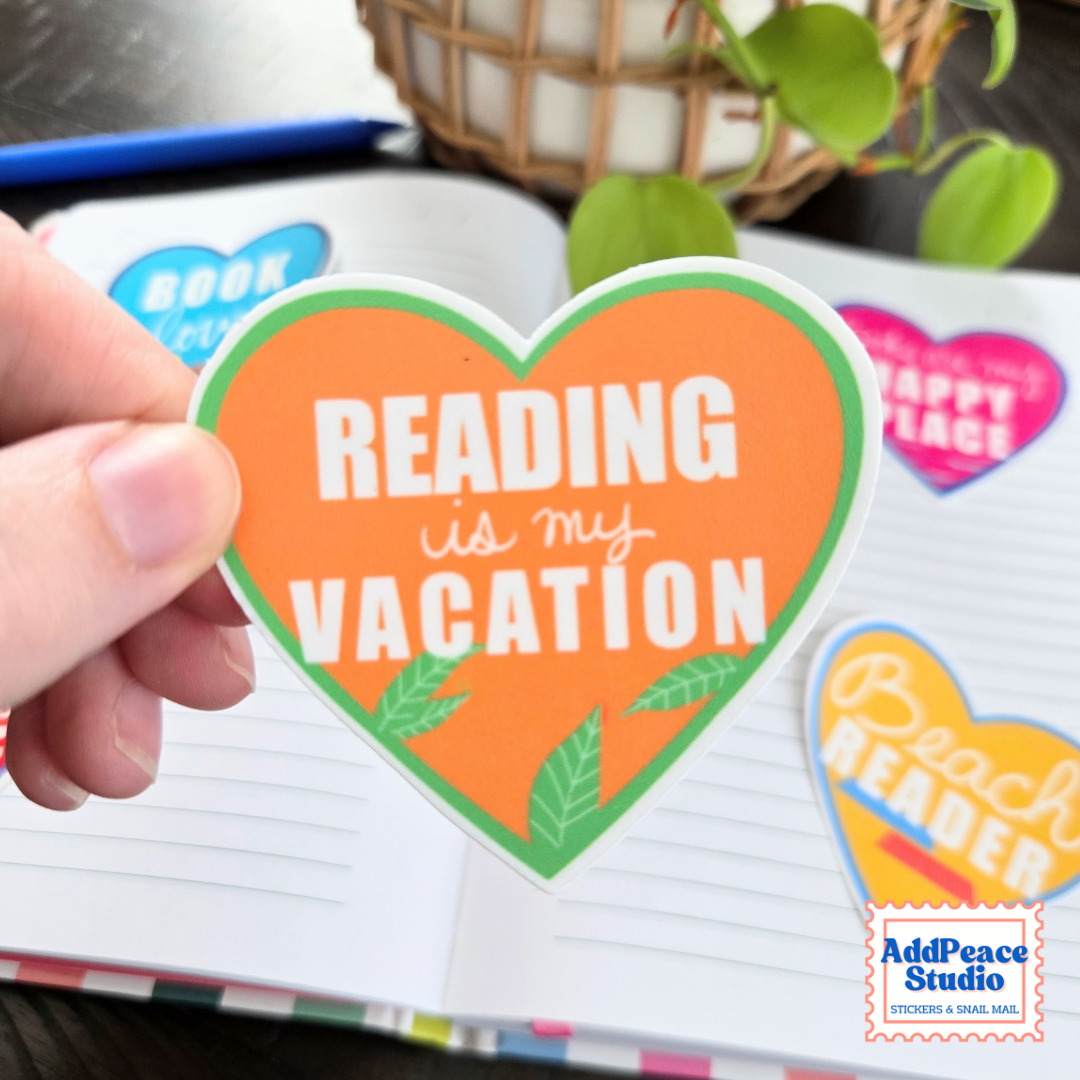 A hand holds a heart-shaped orange sticker that reads 'READING is my VACATION' with green leaves and a green border. The sticker is shown over an open notebook with other colorful stickers, and a plant is in the background.