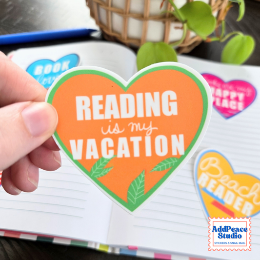 A hand holds a heart-shaped orange sticker that reads 'READING is my VACATION' with green leaves and a green border. The sticker is shown over an open notebook with other colorful stickers, and a plant is in the background.